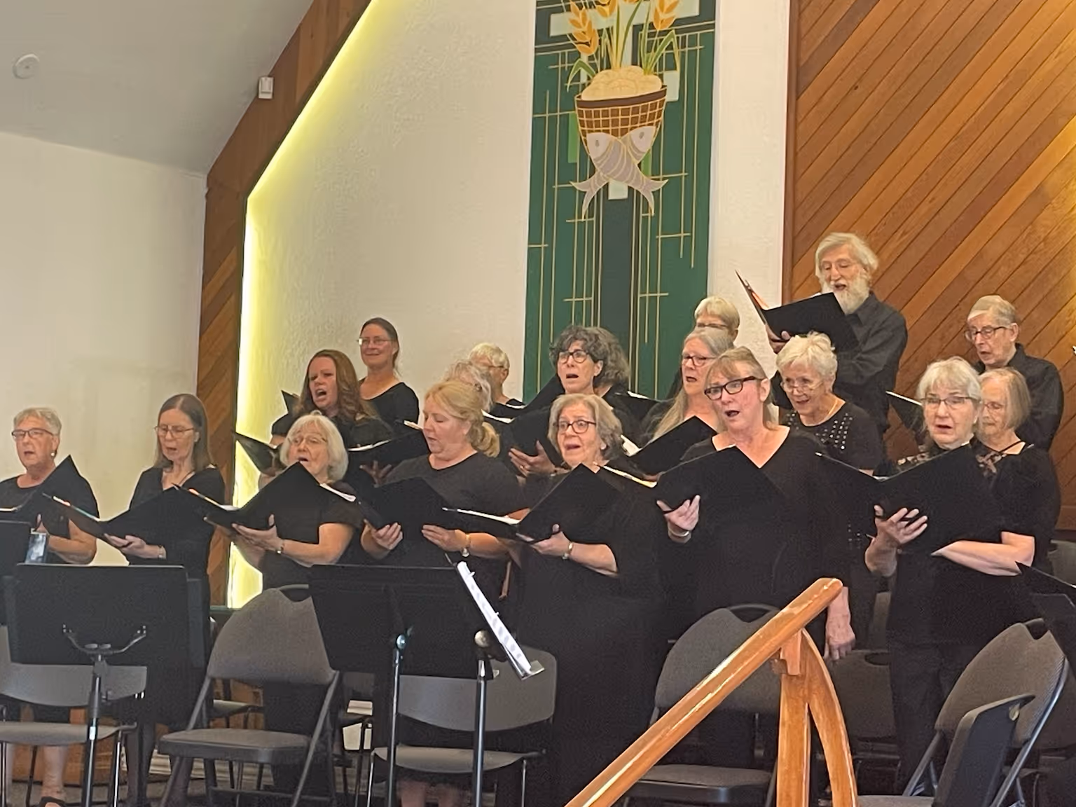 A choir performs during the Classical Finale Concert, standing in rows and singing from black choral folders beneath tall wooden paneling and green hanging banners.
