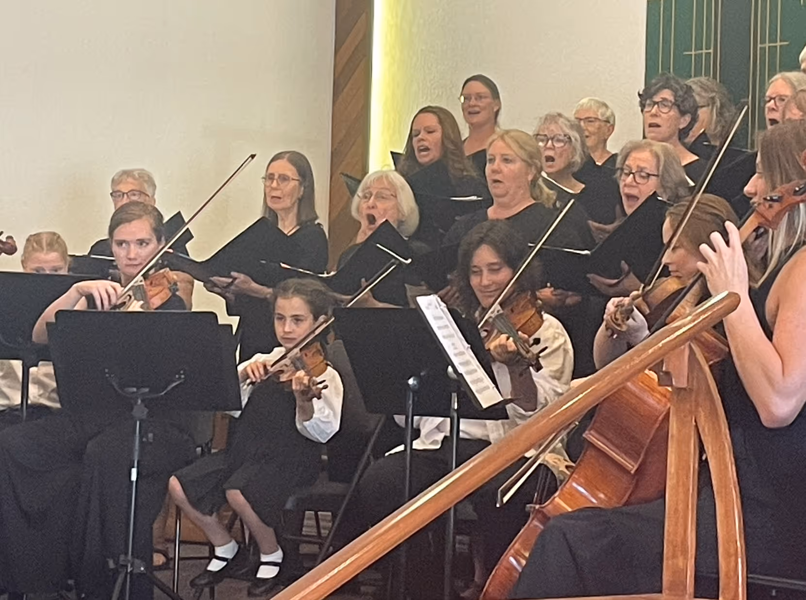 The choir sings alongside a string orchestra during the Classical Finale Concert, with violinists seated in the foreground and singers holding choral folders behind them onstage.