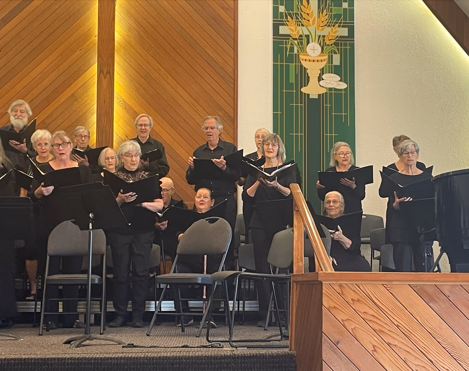 A choir performs during the Classical Finale Concert, with singers standing in rows and holding black choral folders. They sing onstage beneath warm wooden panelling and green hanging banners inside a church setting.