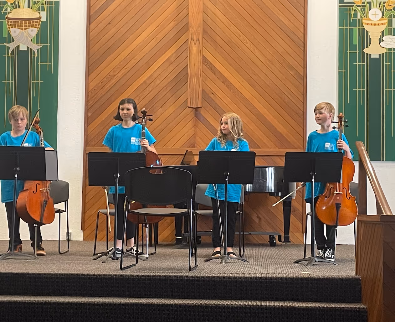 Four classical music students stand onstage with cellos and music stands, wearing matching blue shirts during a student performance. They are positioned in front of a piano and warm wooden paneling inside a concert space.