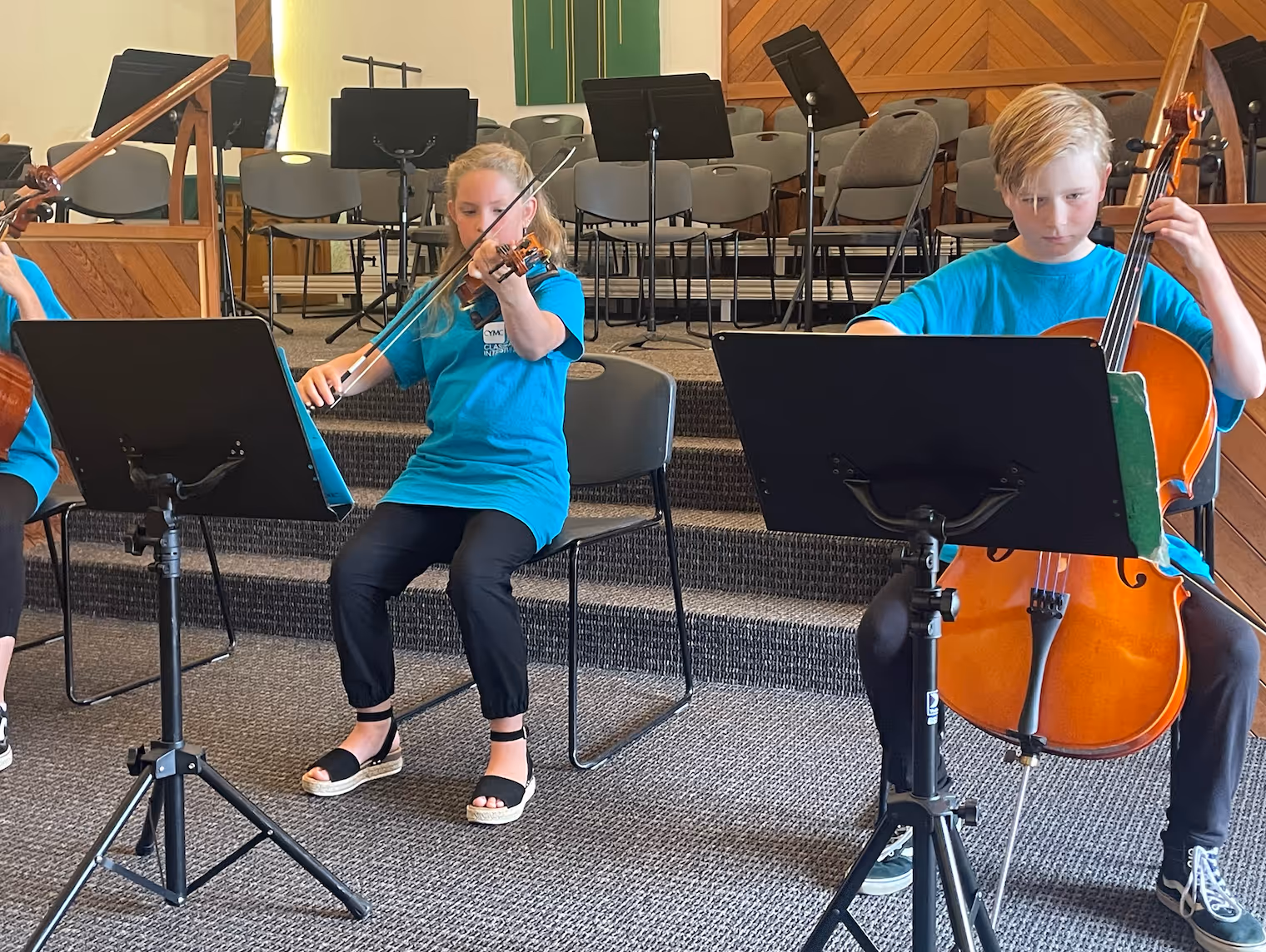 Two young classical string students perform on a church stage, a violinist and a cellist seated side by side, both focused on their sheet music with music stands positioned in front of them.
