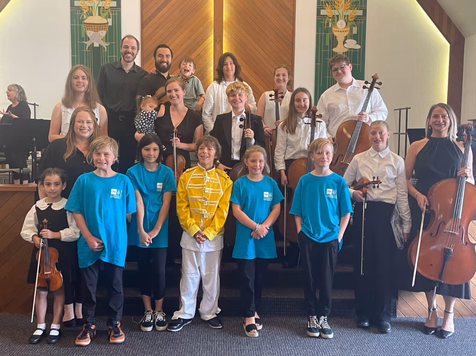 Youth string students stand at the front of a church stage alongside faculty and guest musicians, posing together after a concert. Several students wear bright blue CYMC shirts, one student stands out in a yellow Freddie Mercury–inspired jacket, and others hold violins or cellos. Adults and instructors stand behind them, smiling, emphasizing the intergenerational and community-based nature of the performance.
