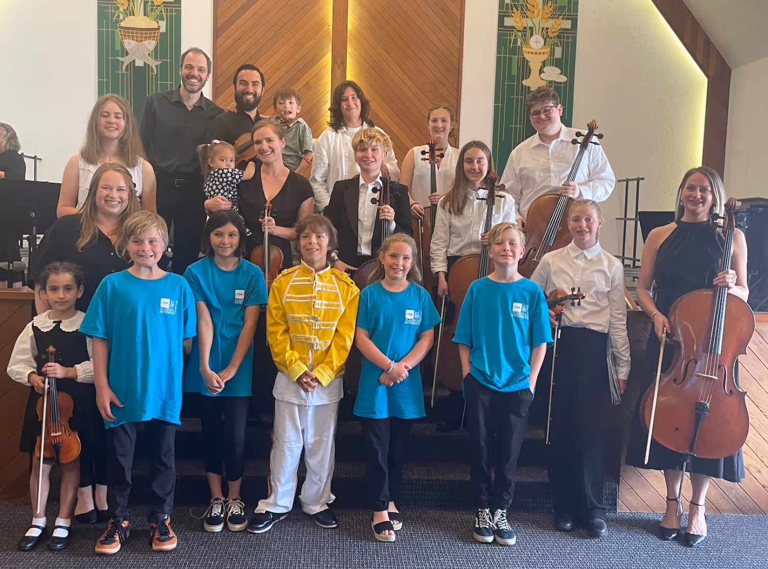 Classical music students and faculty stand together on a church stage following a concert, holding violins, violas, and cellos. Younger students in blue CYMC shirts stand in the front row, while older students and faculty stand behind them. One student in the centre wears a yellow Freddie Mercury–inspired outfit, adding a playful moment to the group portrait.