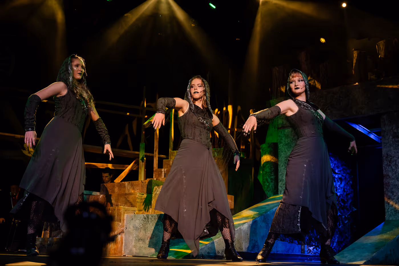 The three Fates appear on stage during Hadestown: Teen Edition, wearing dark, flowing costumes and moving in synchronized poses. Dramatic green and gold lighting and angular set pieces create an ominous, otherworldly atmosphere.