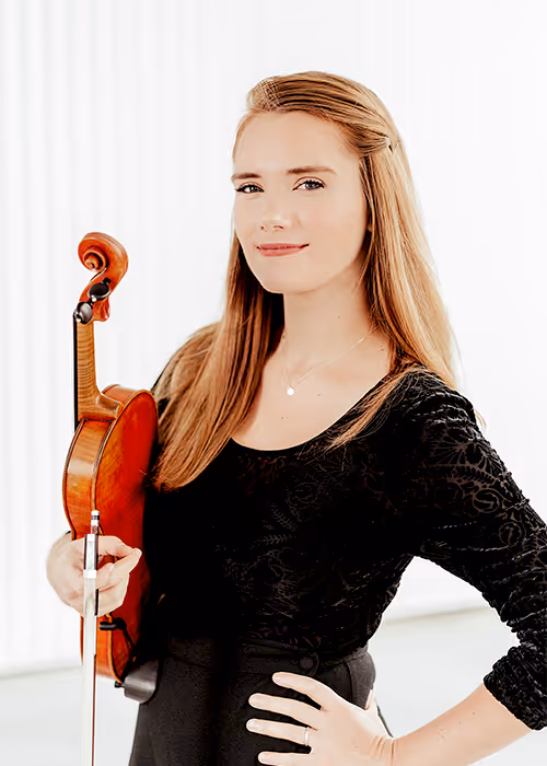 Kathryn Wiebe holds a violin and bow, standing in a light studio setting.