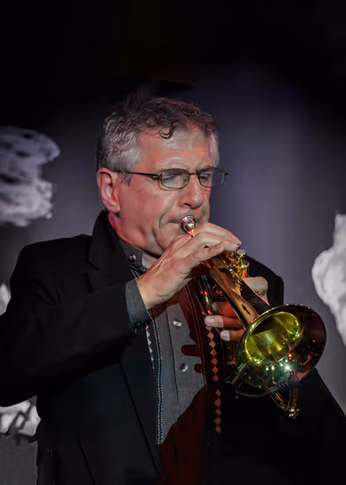 Greg Bush, Jazz Faculty Head, playing trumpet on stage. He wears glasses and a dark jacket, holding a brass trumpet to his lips under dramatic stage lighting with a dark background.