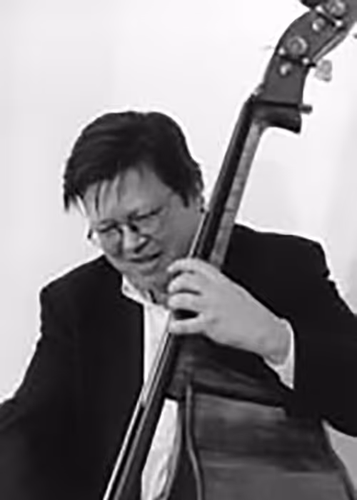 Steve Wallace playing an upright bass, wearing glasses and a dark jacket, focused on the instrument against a plain light background.