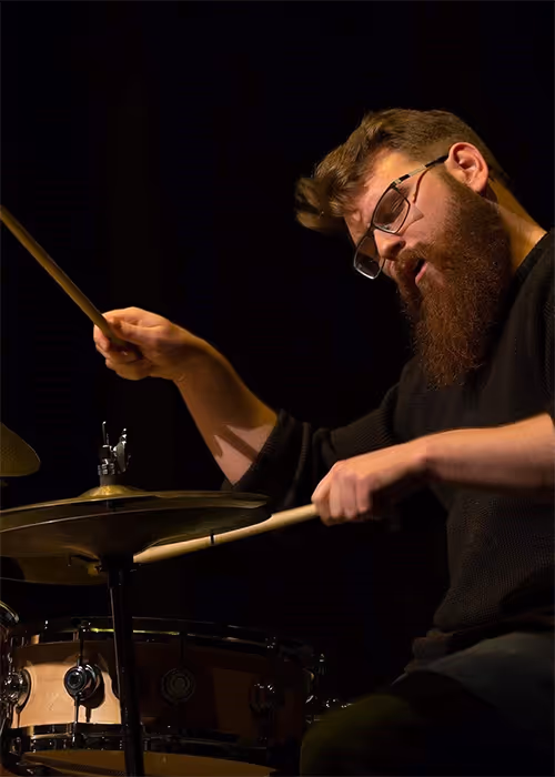 Adam Robertson playing a drum kit on stage, holding drumsticks mid-performance under stage lighting.