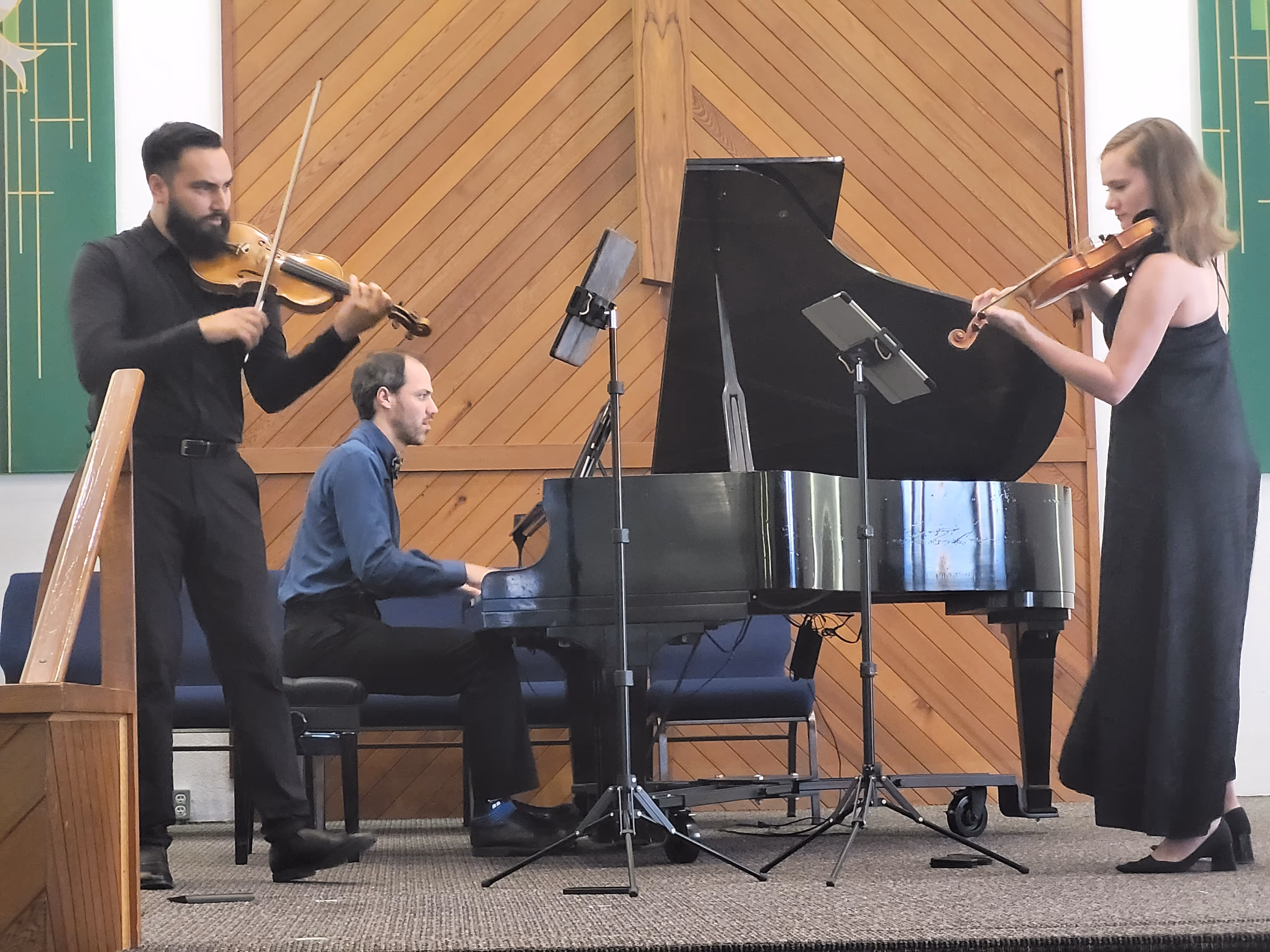 Three musicians perform chamber music in a church setting: a pianist seated at a grand piano in the centre, with two violinists standing on either side, all reading from music stands during a live performance.
