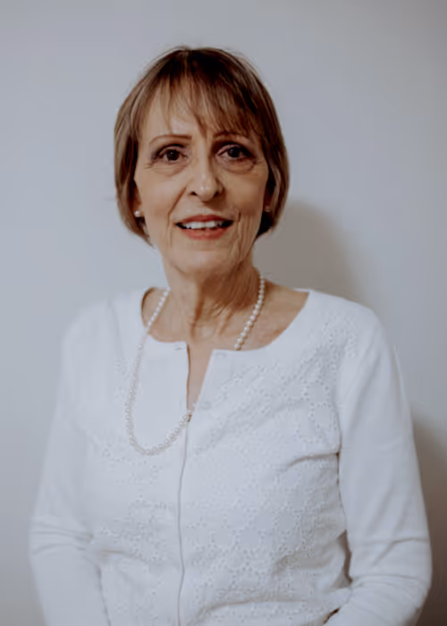 Portrait of Pauline Cote, Vice President of the Comox Valley Youth Music Centre, wearing a white textured cardigan and pearl necklace, standing against a light neutral background.
