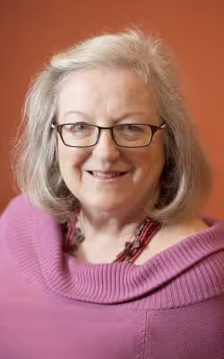 Portrait of Diane Maxted, Director of the Comox Valley Youth Music Centre, smiling and wearing glasses and a pink sweater, posed against a warm, reddish background.