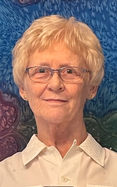 Portrait of Gail Embury, Director of the Comox Valley Youth Music Centre, wearing glasses and a white collared shirt, standing in front of a colorful patterned background and looking directly at the camera.