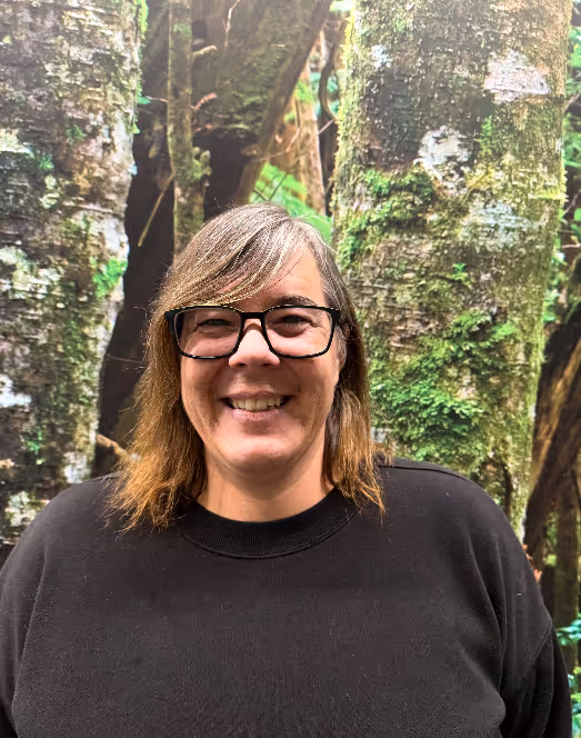 Portrait of Erin Lowdon, Executive Director of the Comox Valley Youth Music Centre, smiling at the camera while standing outdoors in front of moss-covered trees, wearing glasses and a black sweater.