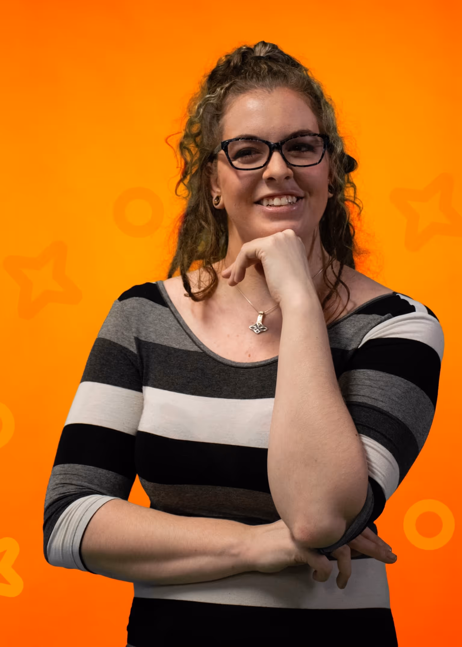 Portrait of Tara Dawn Jakubiec, Graphic Designer and Registrar, wearing glasses and a striped top, smiling confidently with her hand resting on her chin against a bright orange background.