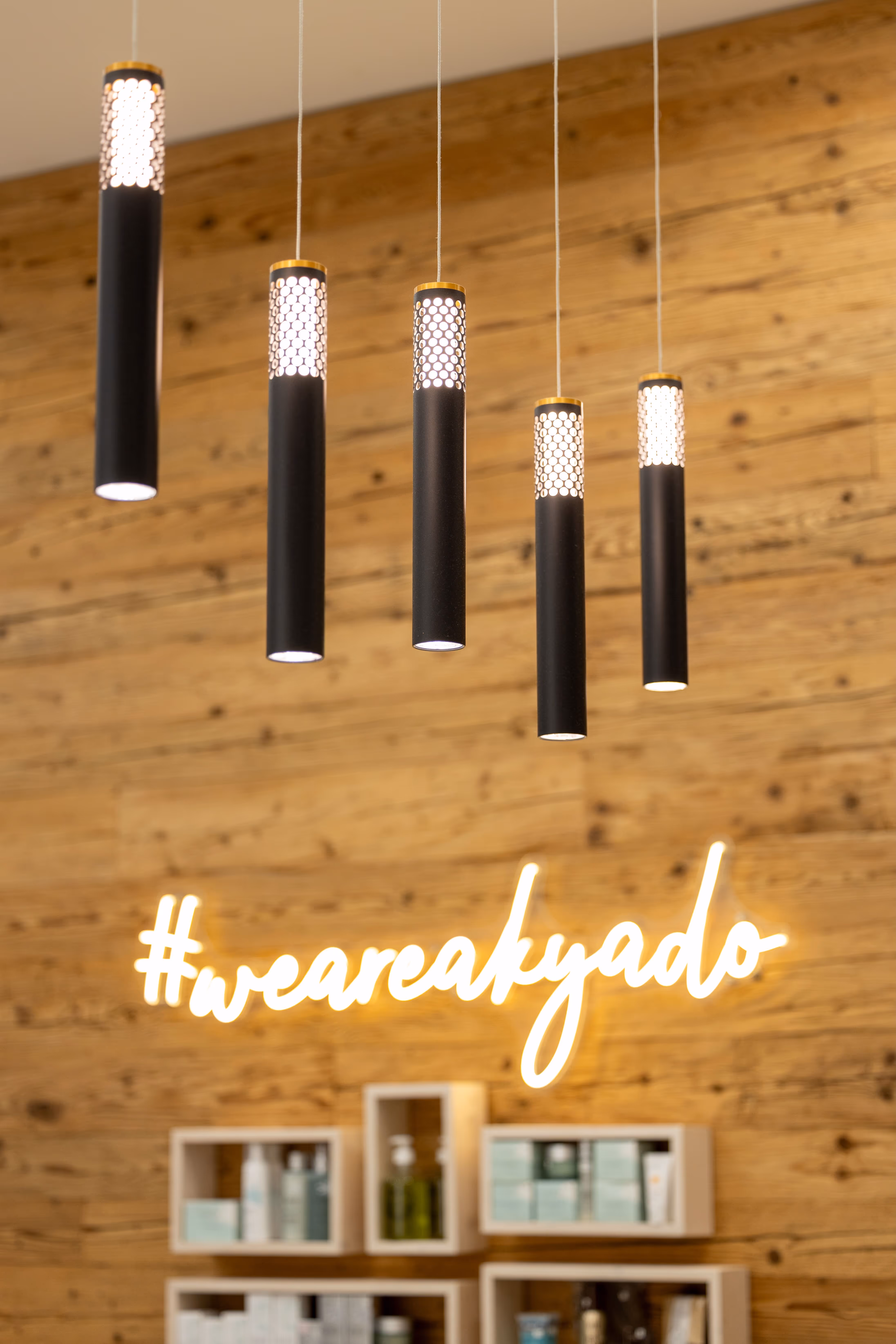 Cinq suspensions noires cylindriques avec lumière blanche au sommet, devant un mur en bois avec le lettrage lumineux #weareakyado.