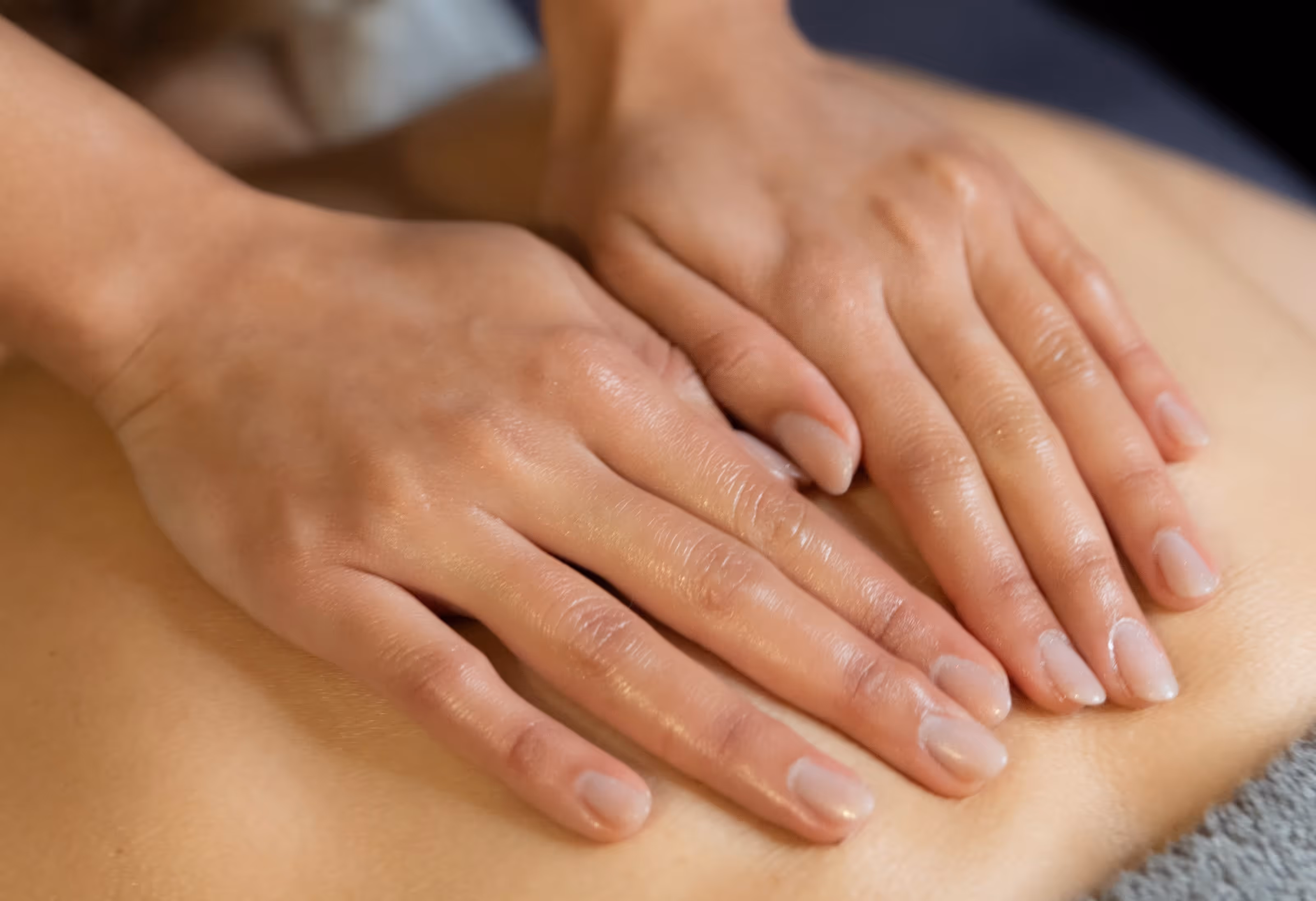 Vue rapprochée de mains de praticienne effectuant un massage relaxant sur le dos d'une personne avec de l'huile de soin, dans une ambiance douce.