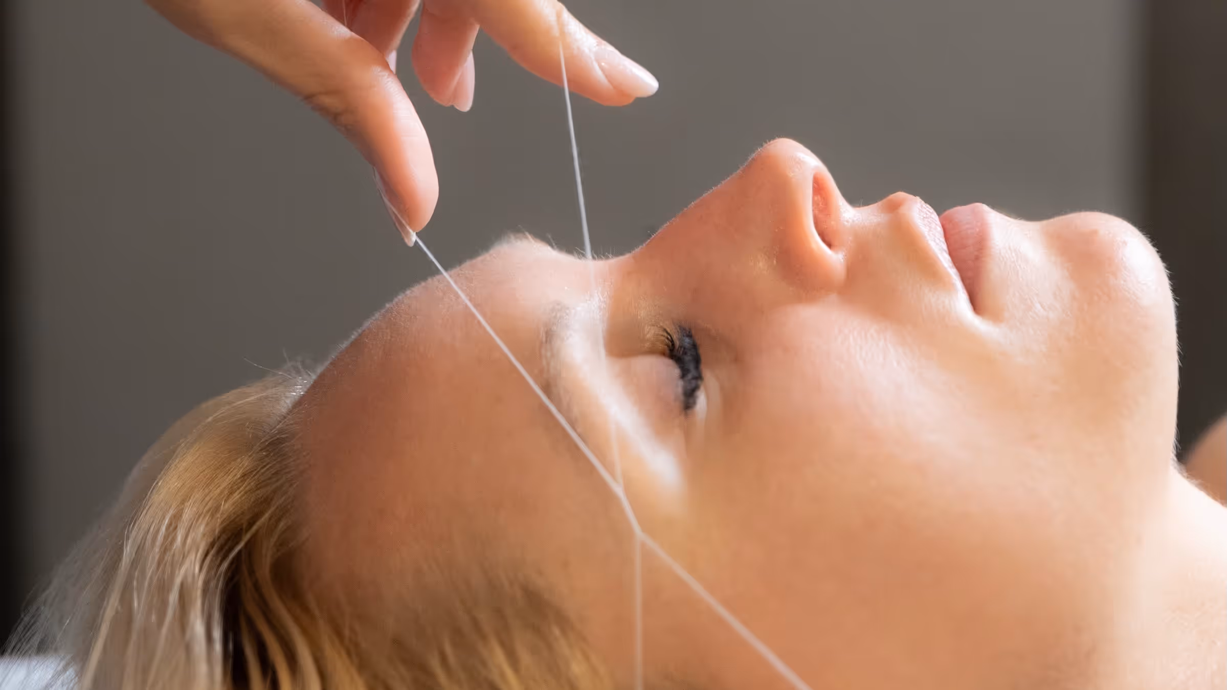 Gros plan sur une séance d'épilation au fil des sourcils pratiquée sur une cliente aux yeux fermés dans un institut de beauté.