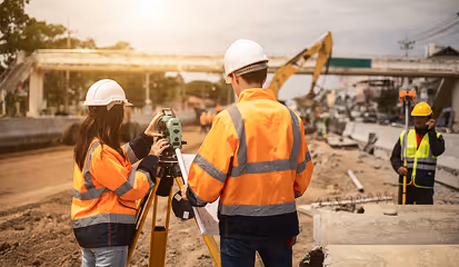 Two construction workers in orange safety jackets using surveying equipment at a roadwork site while a third worker stands near a measuring device.