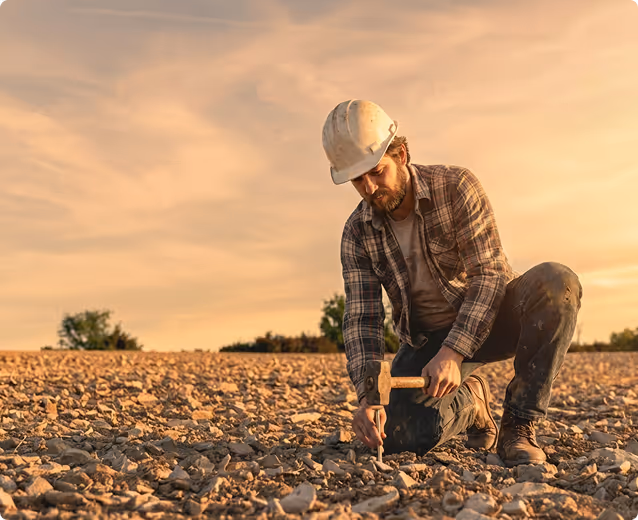Man wearing a hard hat and plaid shirt kneeling on rocky ground, hammering a stake into the soil at sunset.