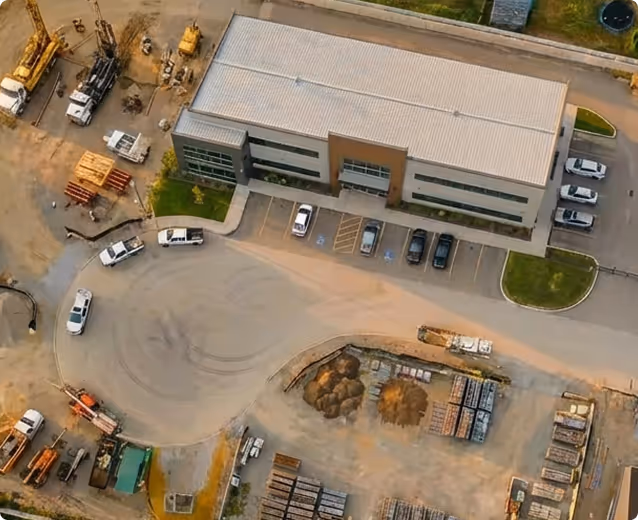 Aerial view of a modern industrial building with parking lot and construction equipment nearby on a dirt lot.