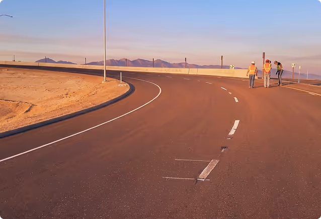 Three construction workers on an empty curved road near a desert with mountains in the distance.