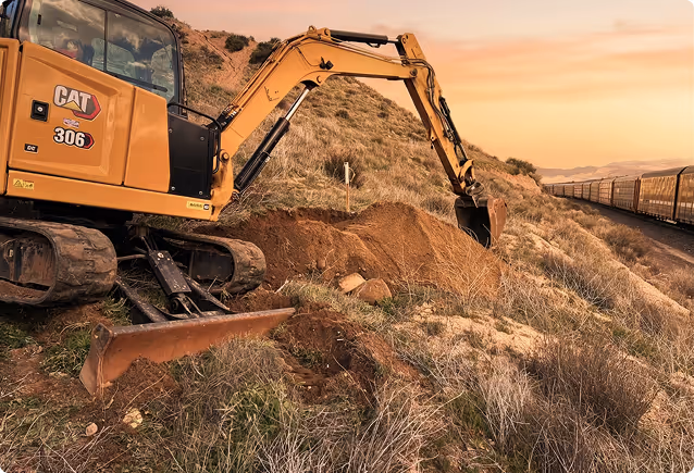 Yellow CAT 306 excavator digging earth on a hillside near a train in sunset light.