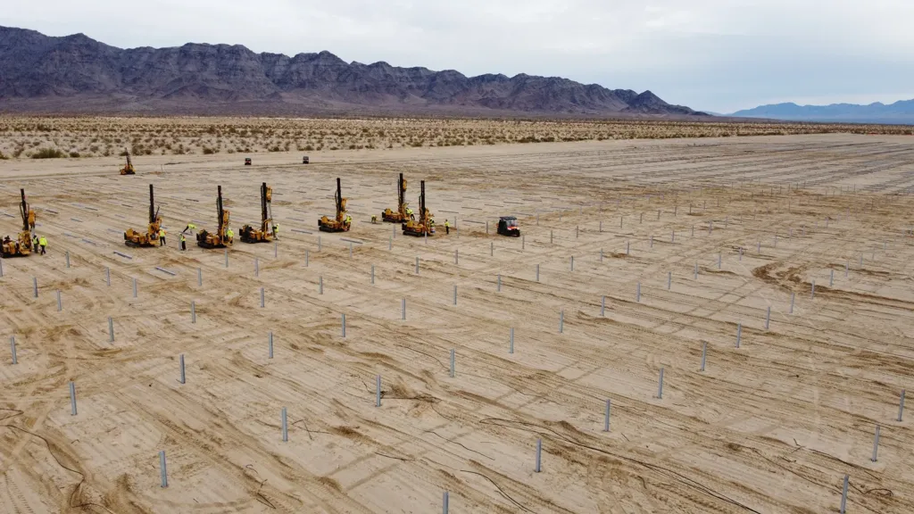 Construction site with machinery and workers installing metal posts in a desert landscape with mountains.