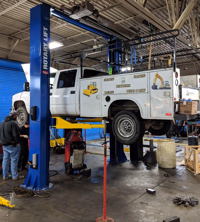 White service truck elevated on a blue Rotary Lift inside a garage with workers nearby and equipment on the floor.