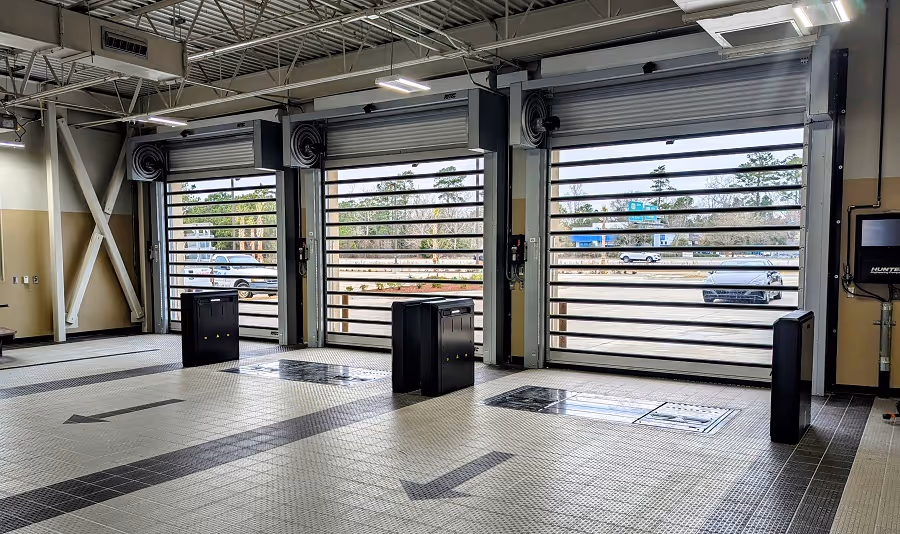 Interior of a car service bay with three open garage doors, tiled floor with directional arrows, and wheel alignment equipment.