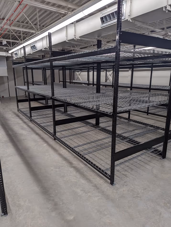Empty black metal wire shelving units in a large industrial storage room with concrete floor and exposed ceiling ducts.