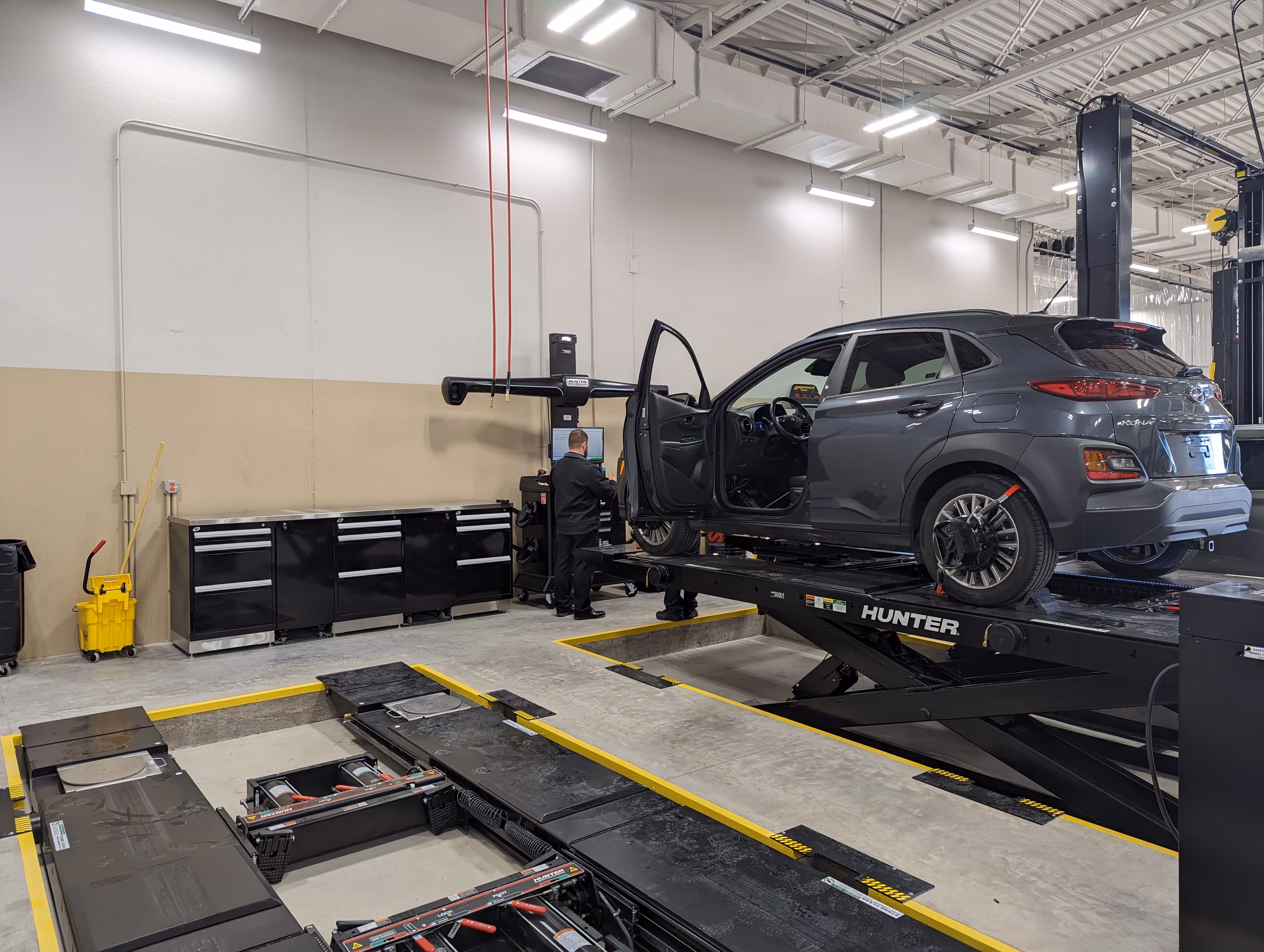 Gray Hyundai Kona lifted on a Hunter alignment machine with a technician checking a computer in a clean automotive service garage.