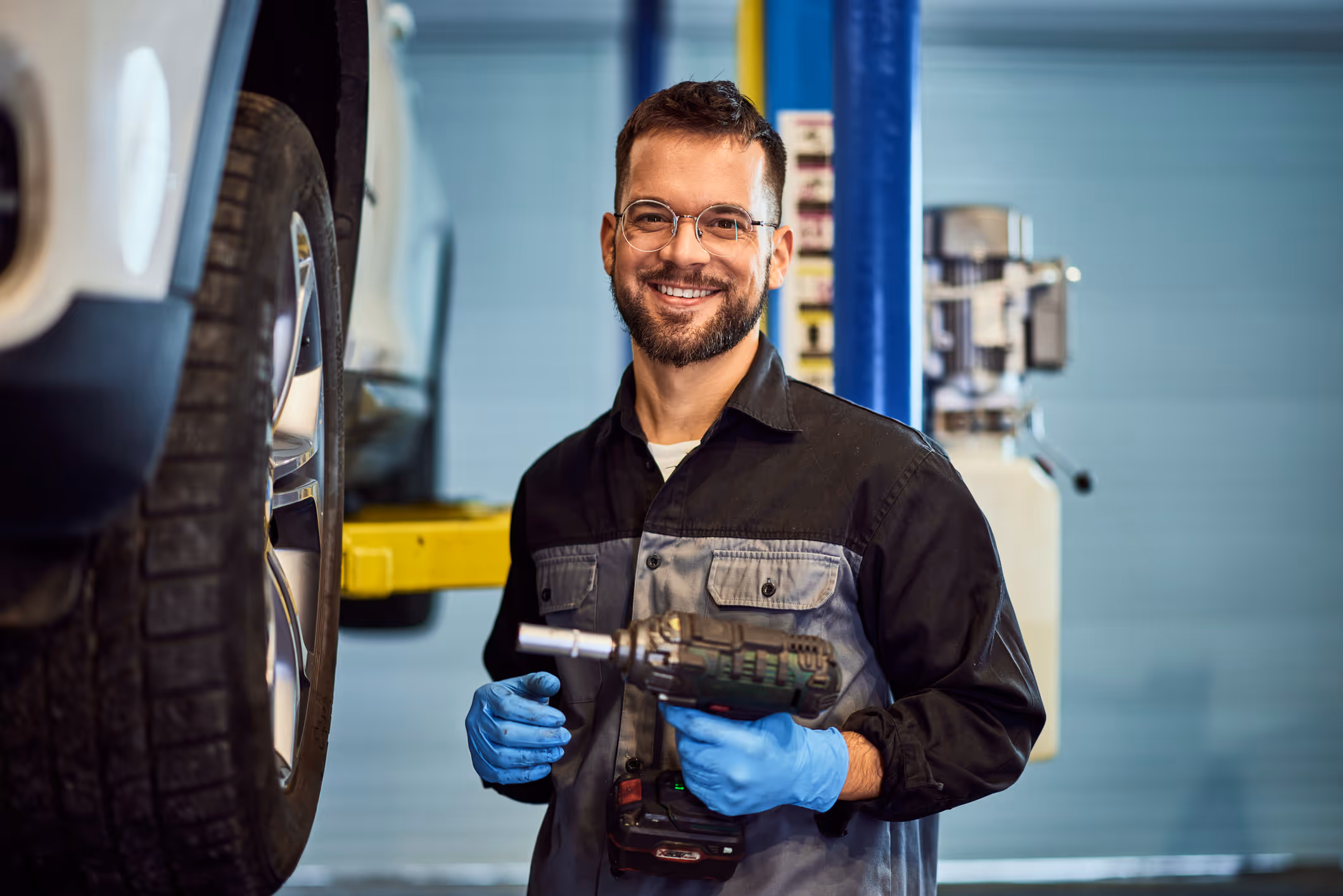 Smiling mechanic wearing glasses and blue gloves holding a power tool near a vehicle tire in a garage.