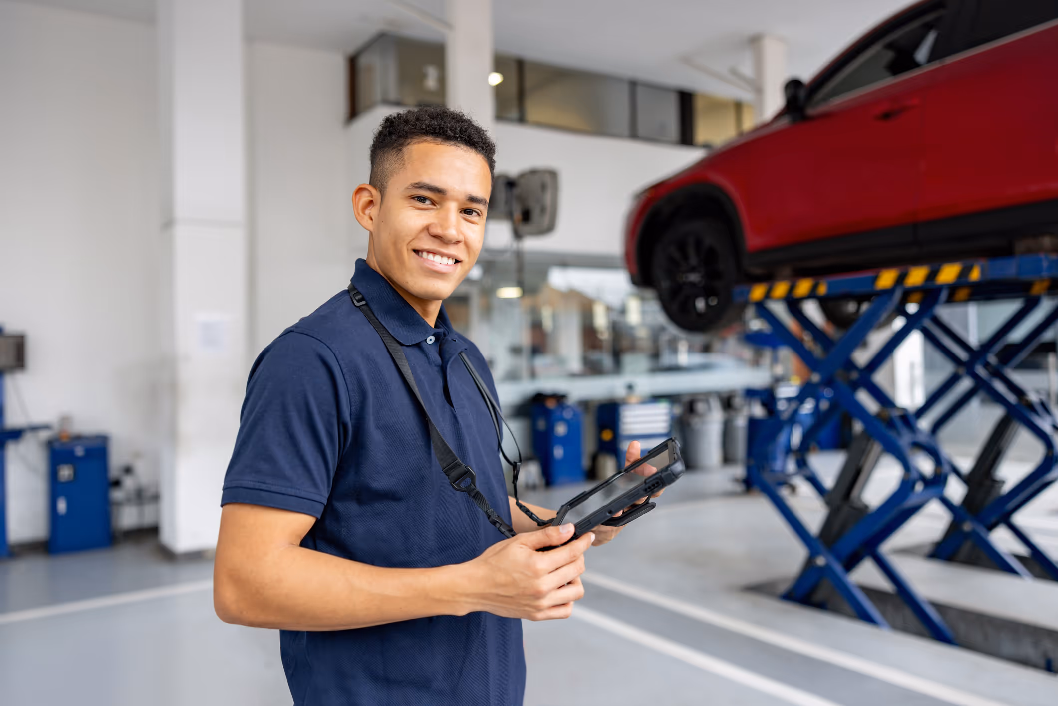 Smiling mechanic in a navy shirt holding a tablet in an auto repair shop with a red car lifted on a hydraulic lift.