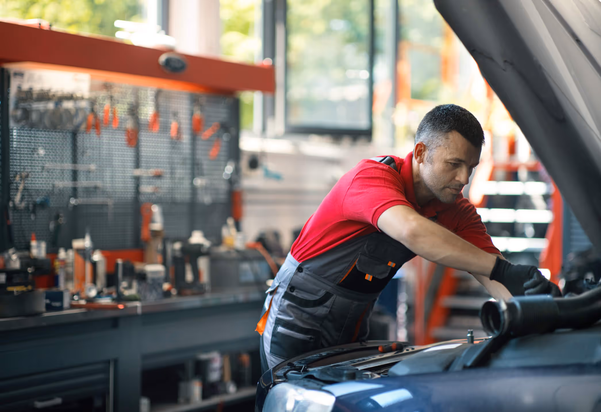 Mechanic in a red shirt and gray overalls working on a car engine with the hood open in a workshop.
