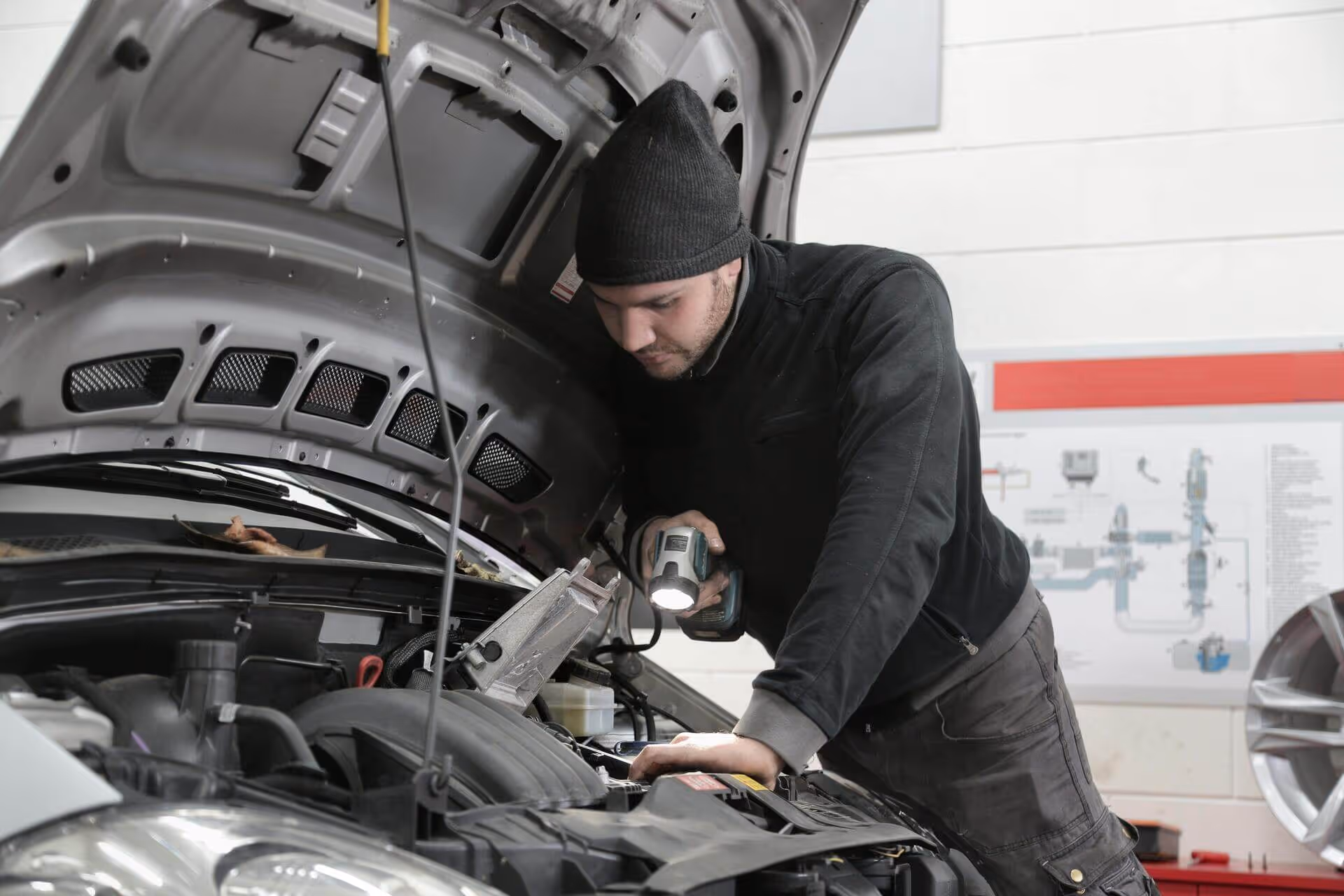 Mechanic wearing a black beanie and jacket inspecting a car engine with a flashlight in a garage.
