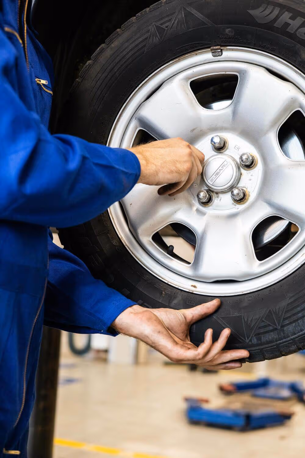 Mechanic in blue coveralls inspecting a car tire with silver alloy wheel in an auto repair shop.