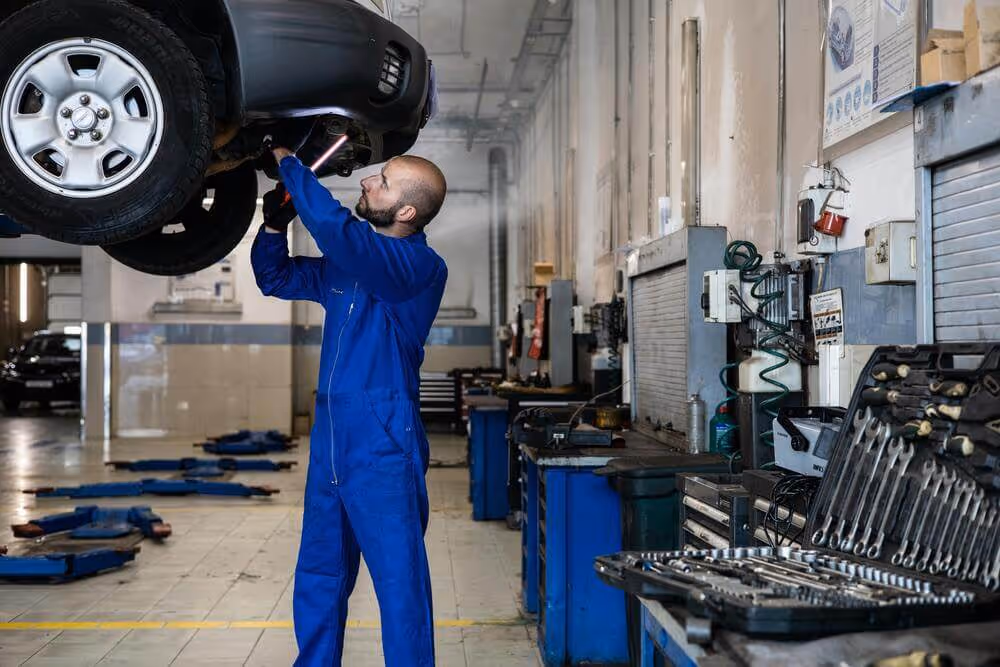 Mechanic in blue coveralls inspecting underside of a raised car in a spacious auto repair shop with tools and equipment visible.