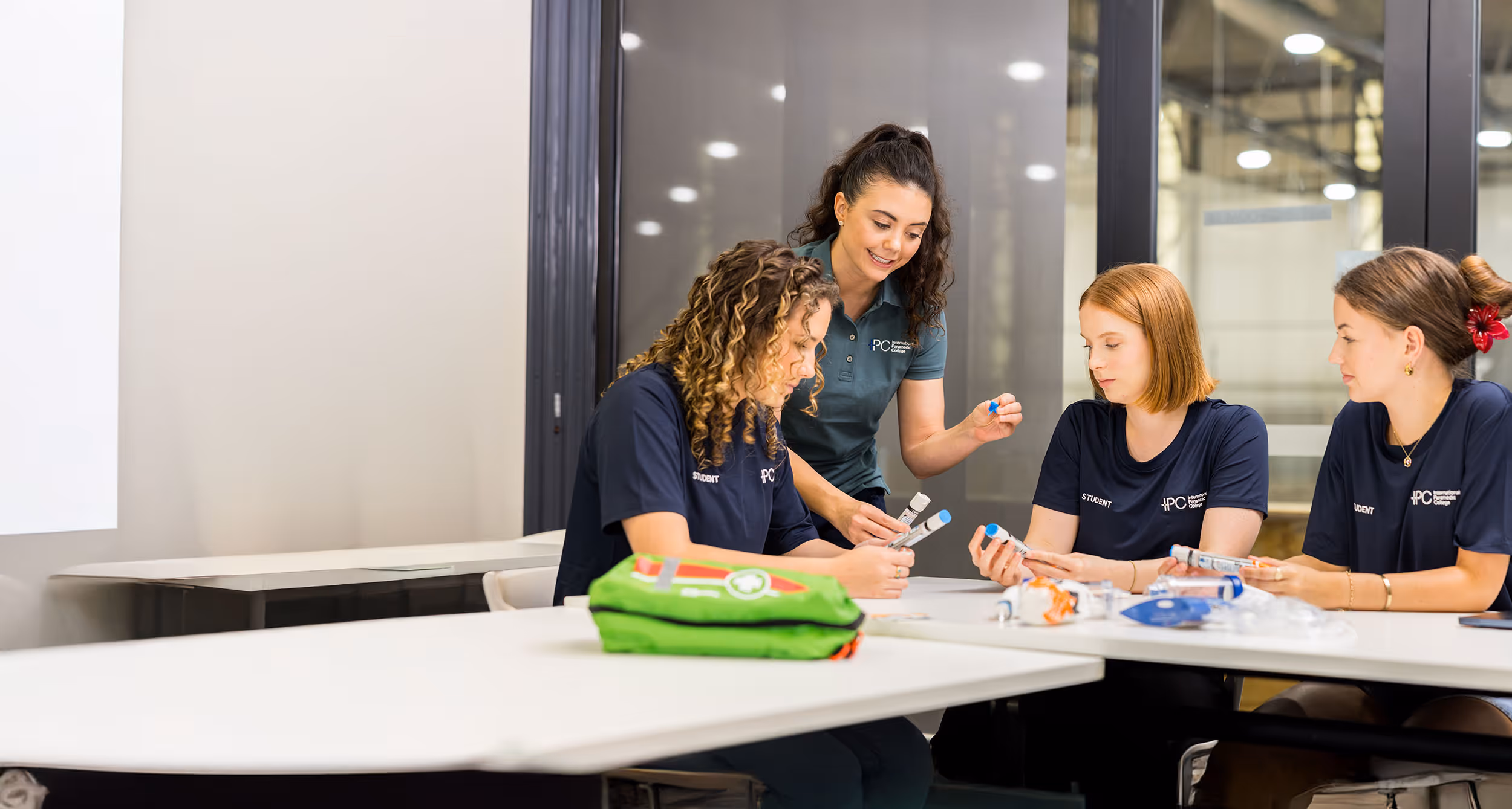 Four women in navy blue shirts with 'PC' logo study medical equipment tubes around a table in a bright classroom.