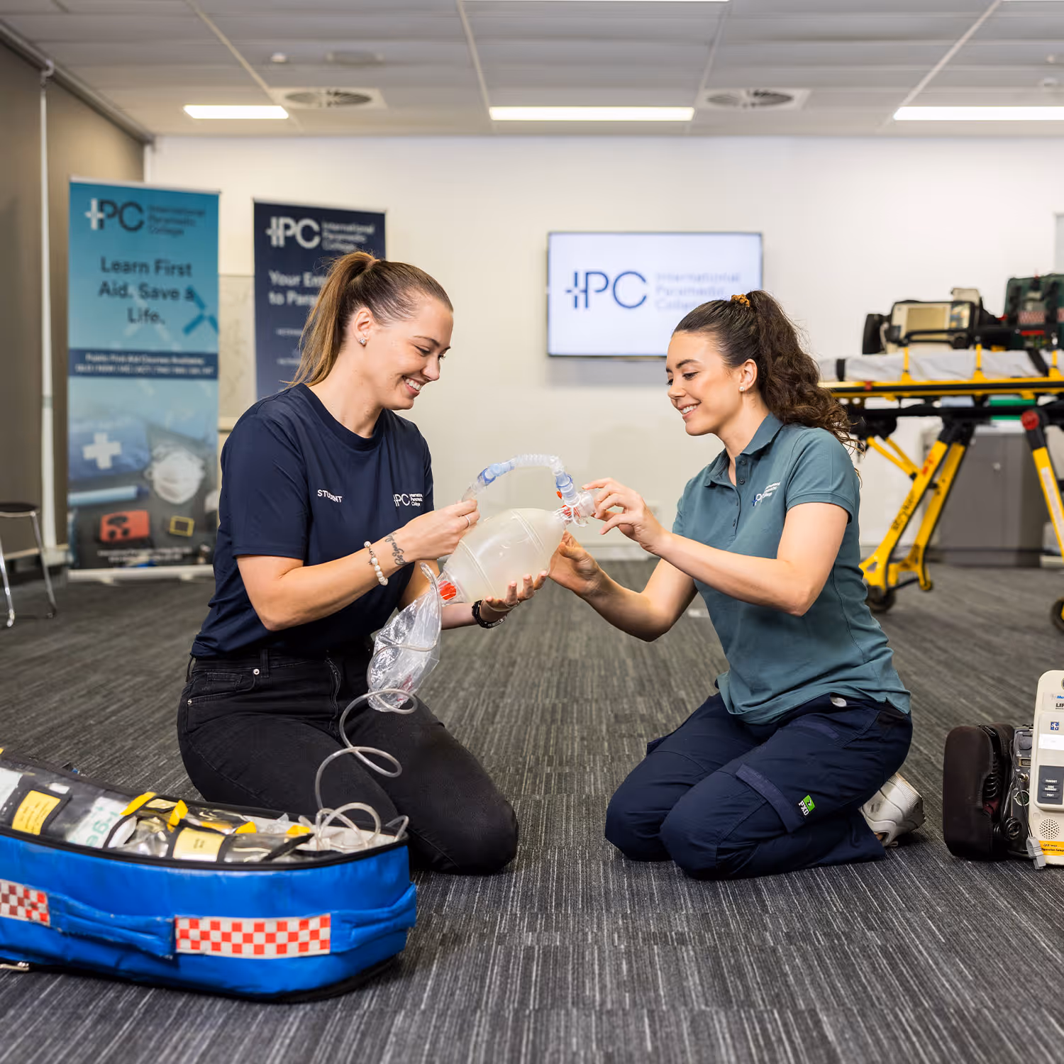 Two women kneeling on the floor, practicing with a manual resuscitator bag in a training room with medical equipment.