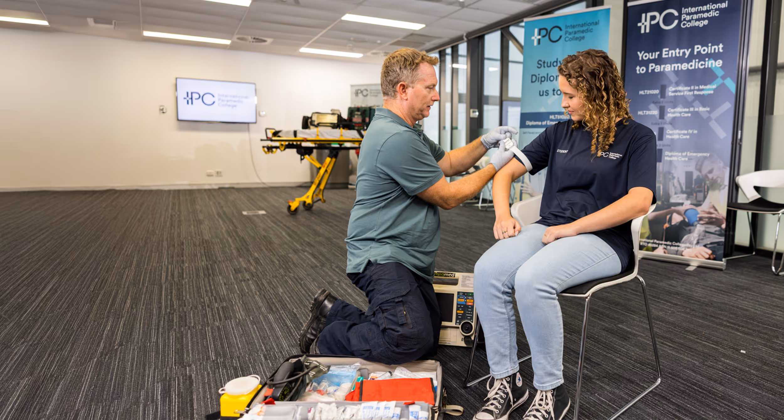 A paramedic instructor kneels applying a tourniquet to a student’s arm in a training room with medical equipment and banners in the background.