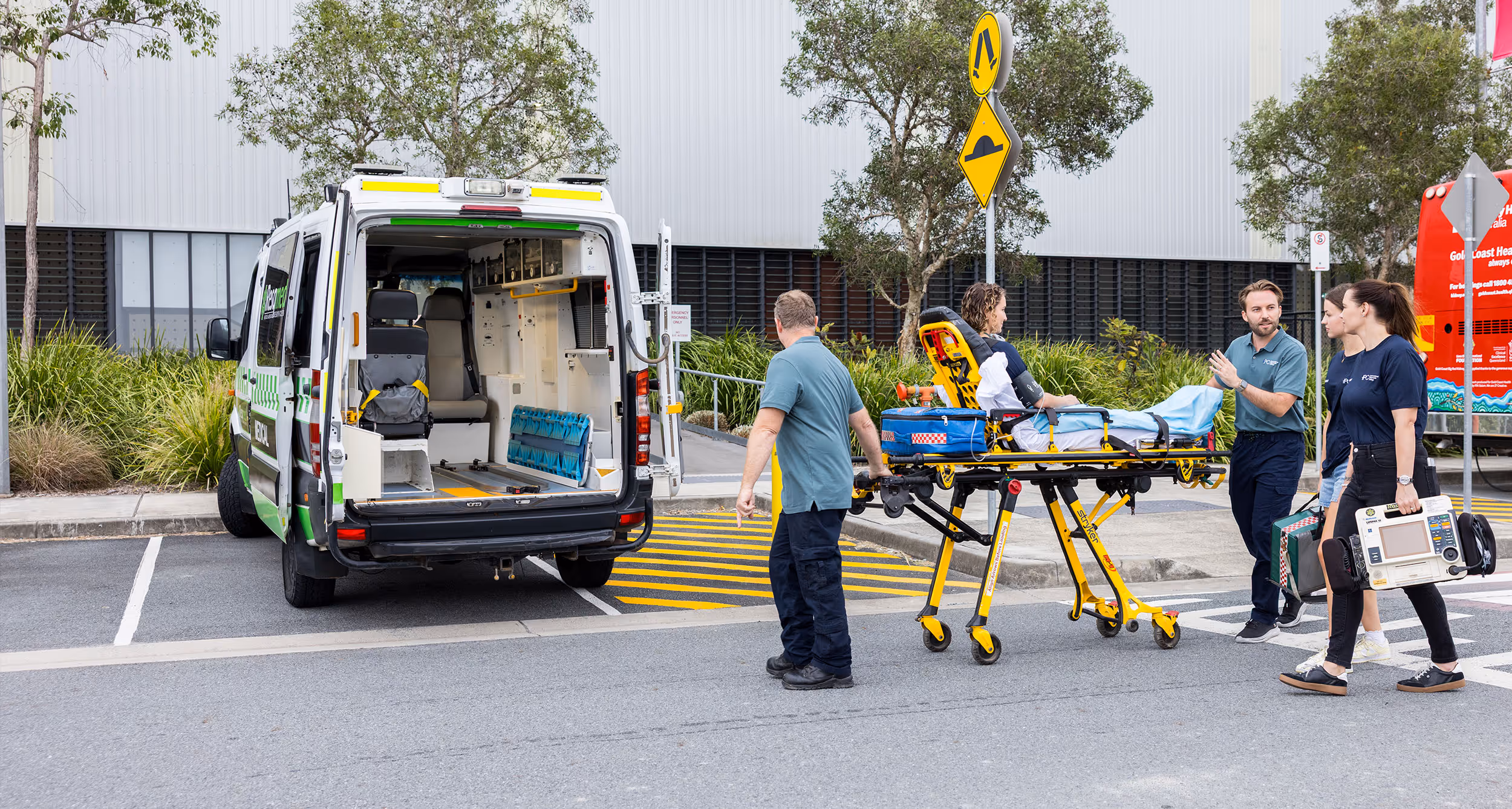 Paramedics transporting a patient on a stretcher towards an open ambulance in an outdoor urban area.