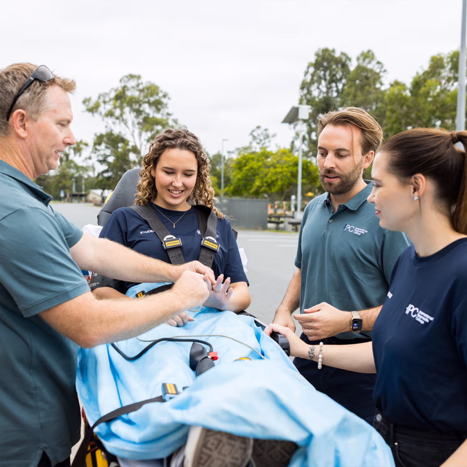 Three paramedics attending a female patient lying on a stretcher covered with a blue blanket outdoors.