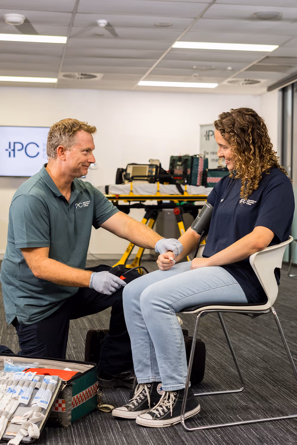 Male instructor kneeling and checking the blood pressure of a seated female student in a medical training setting.