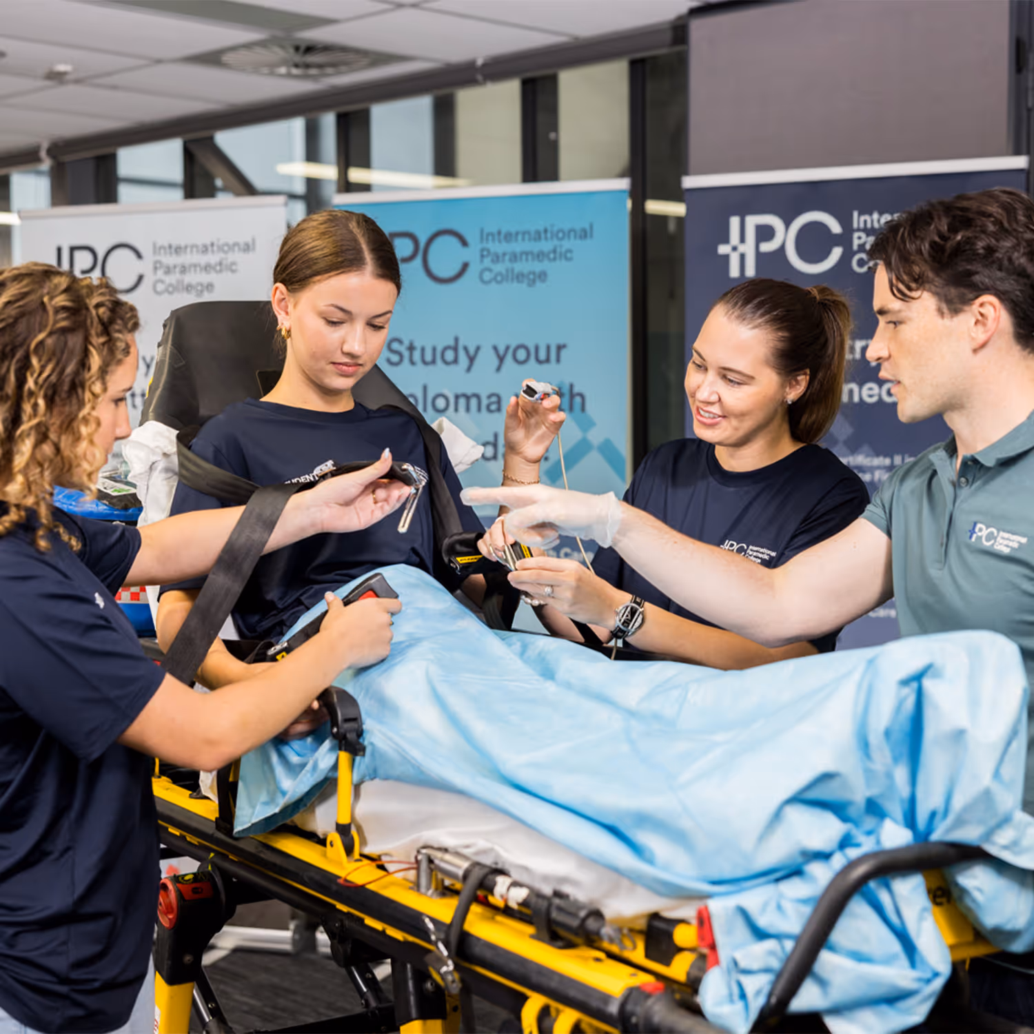 Four individuals in International Paramedic College shirts practicing patient care on a stretcher with medical equipment.