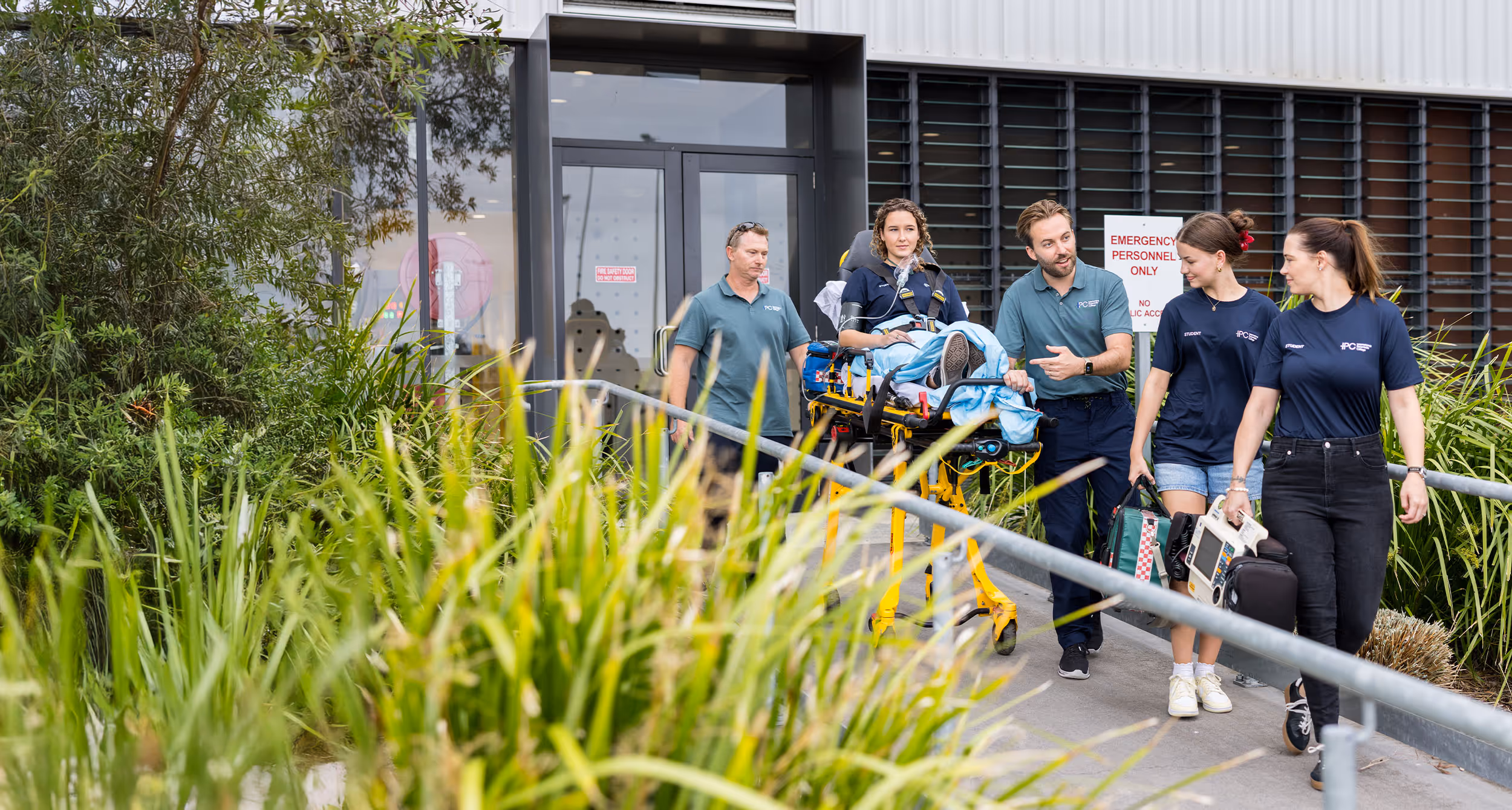 Paramedics training with a patient on a stretcher outside a medical facility, carrying emergency equipment.