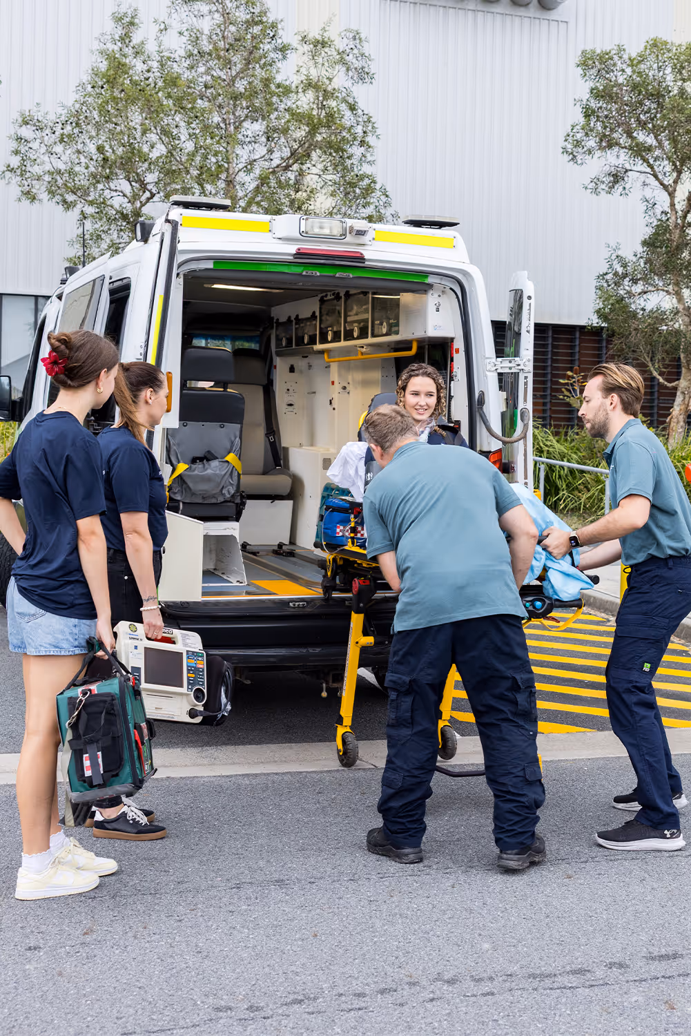 Paramedics loading a smiling patient on a stretcher into an ambulance while two other medical staff hold emergency equipment nearby.
