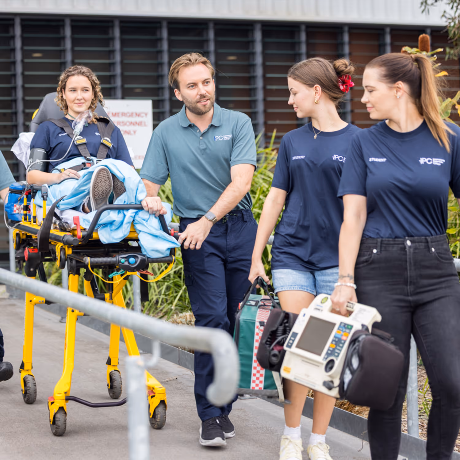 Paramedic pushing a stretcher with a patient while two students carry medical equipment walking alongside.