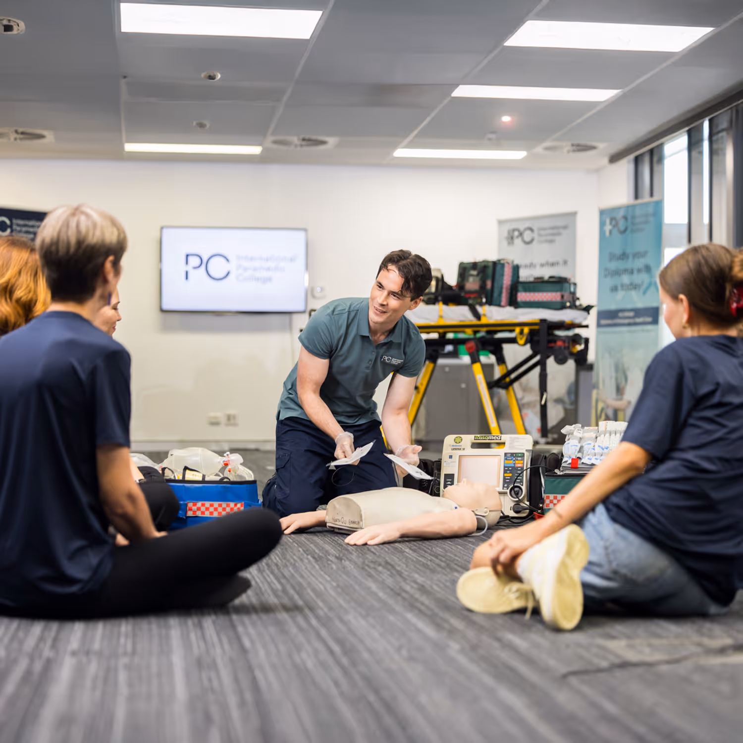 Instructor demonstrating CPR with defibrillator pads on a training manikin to seated students in a classroom.