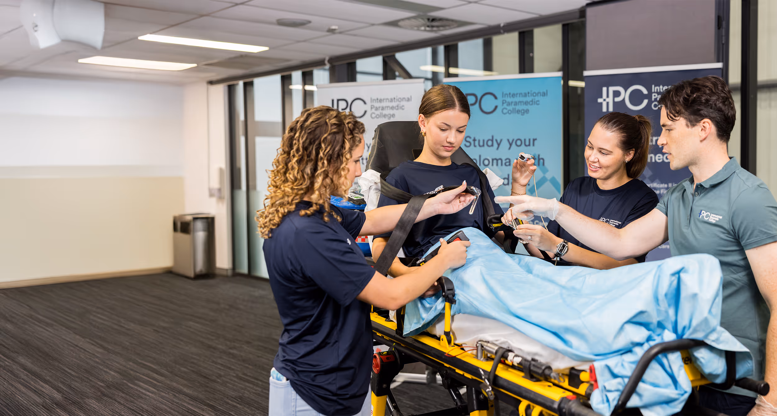 Four paramedic students practicing patient care on a stretcher in a clinical classroom setting.