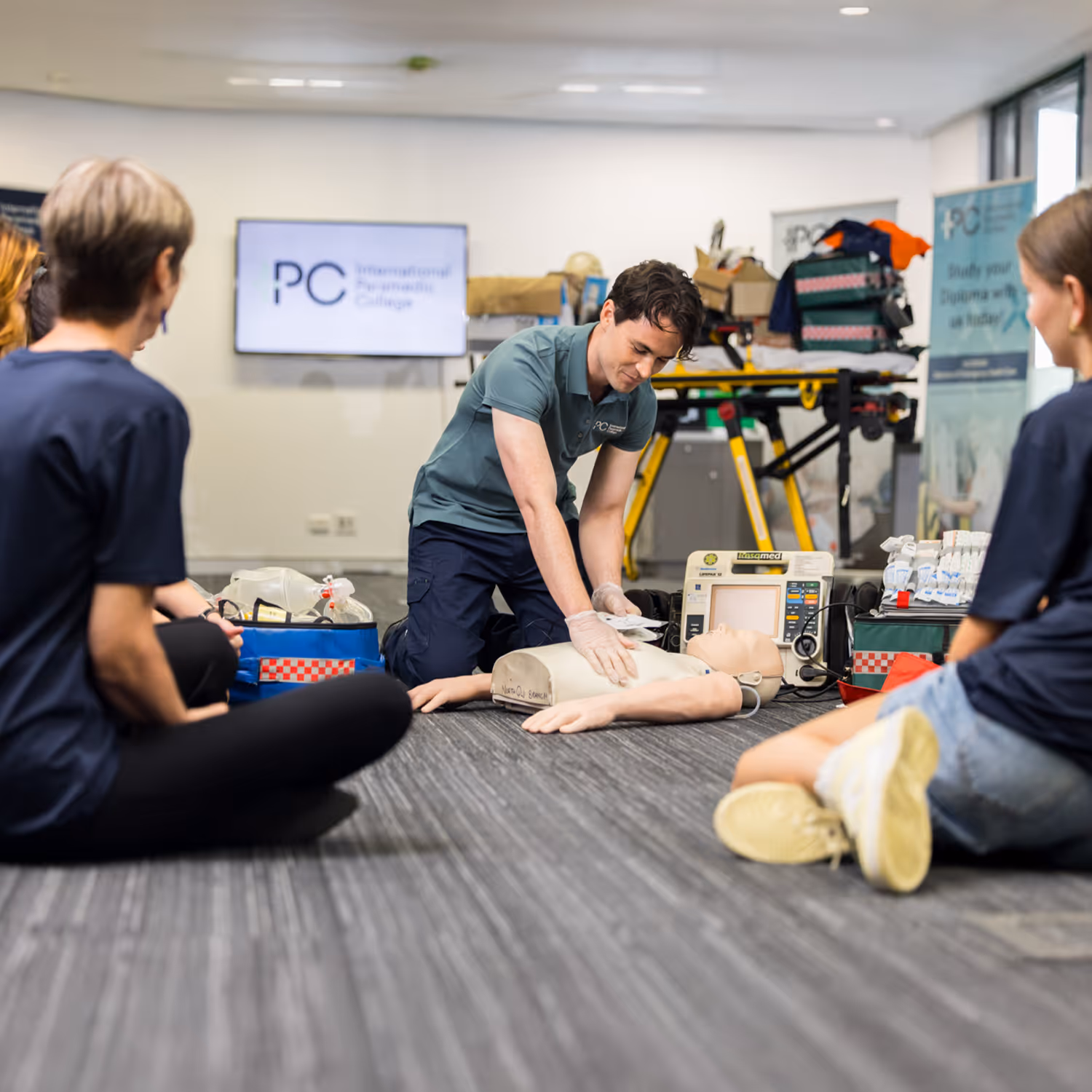 Instructor demonstrating CPR on a training mannequin to seated students in a classroom.